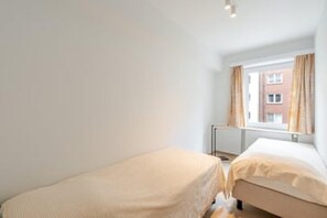 A room with two beds, a window with curtains, and a radiator. - Blankenberge Apartment for 4 (Blankenberge)
