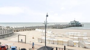 Un hotel frente a la playa con un muelle que se adentra en el mar, una torre de salvavidas y sombrillas de playa.