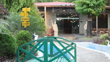 Un banco metálico verde en un jardín con un letrero que indica la cercanía de una piscina y un restaurante.