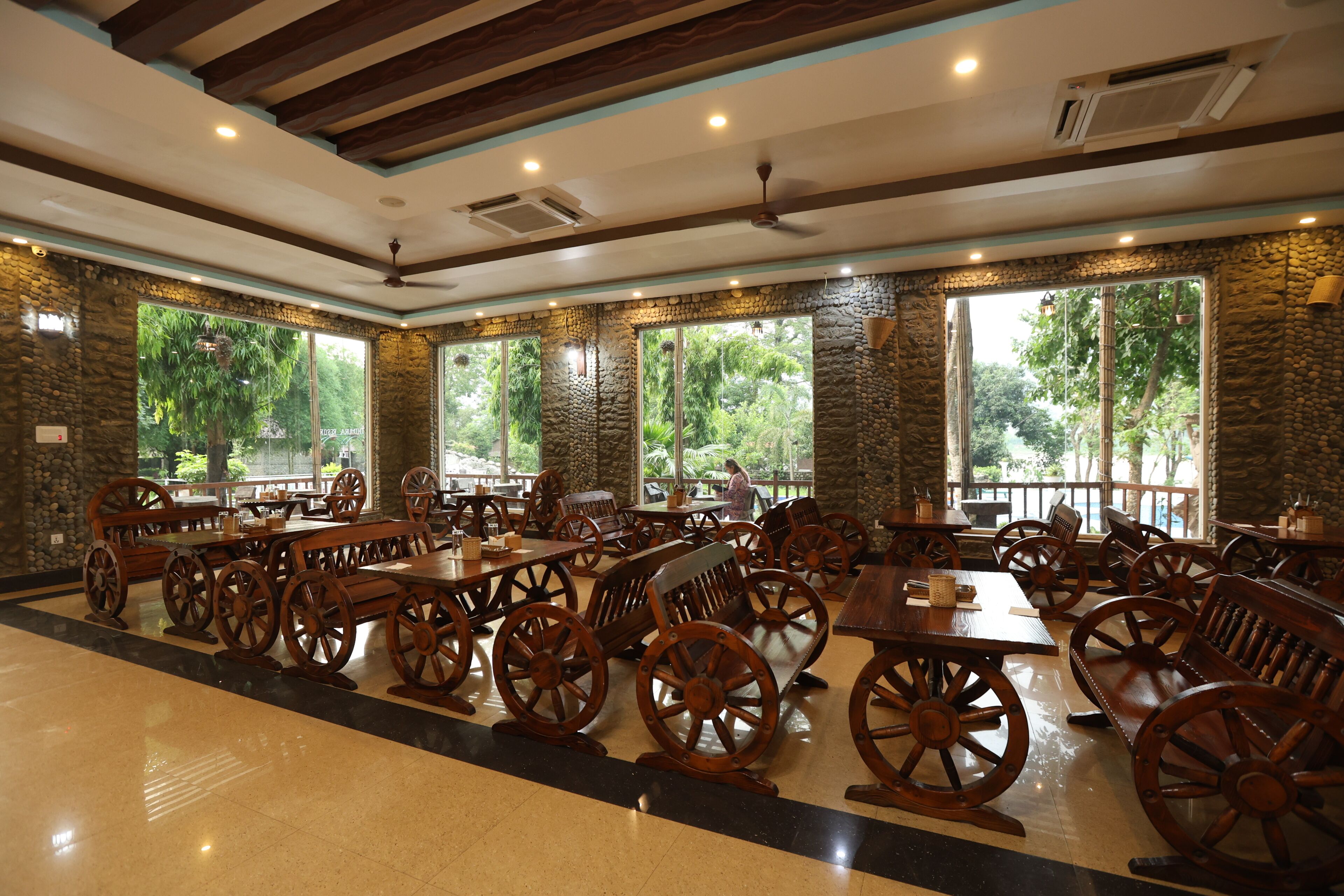Un restaurante con paredes de piedra, grandes ventanales, mesas de madera con ruedas y piso de baldosas.