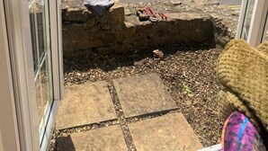 Un edificio de piedra con techo de tejas, un coche blanco estacionado en la calle y una pared de piedra con una pequeña abertura.