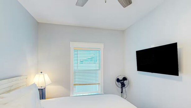A bedroom with a bed, a bedside lamp, a window with blinds, a wall-mounted TV, and a standing fan. - Days at the Beach | Pet Friendly | PCB (Panama City Beach)