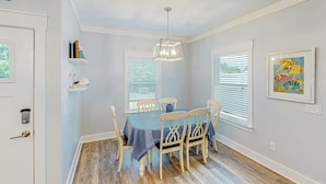 A dining area with a table set for four, wooden chairs, a chandelier, and two windows with blinds. - Days at the Beach | Pet Friendly | PCB (Panama City Beach)