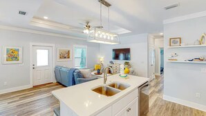 A modern kitchen with a central island, a dining area with a blue sofa, and a ceiling fan. - Days at the Beach | Pet Friendly | PCB (Panama City Beach)
