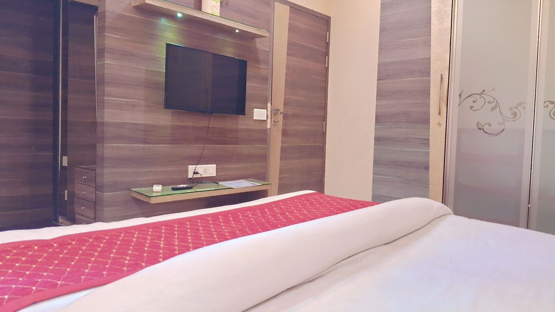 A hotel room with a bed, a flat-screen TV mounted on the wall, a small table with a remote control, and a wardrobe with a mirror.
