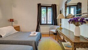 Ein Hotelzimmer mit zwei Betten, einem fenster mit Vorhängen, einem Nachttisch mit Lampe, einem Sessel, einem Spiegel und einer Vase mit Blumen.