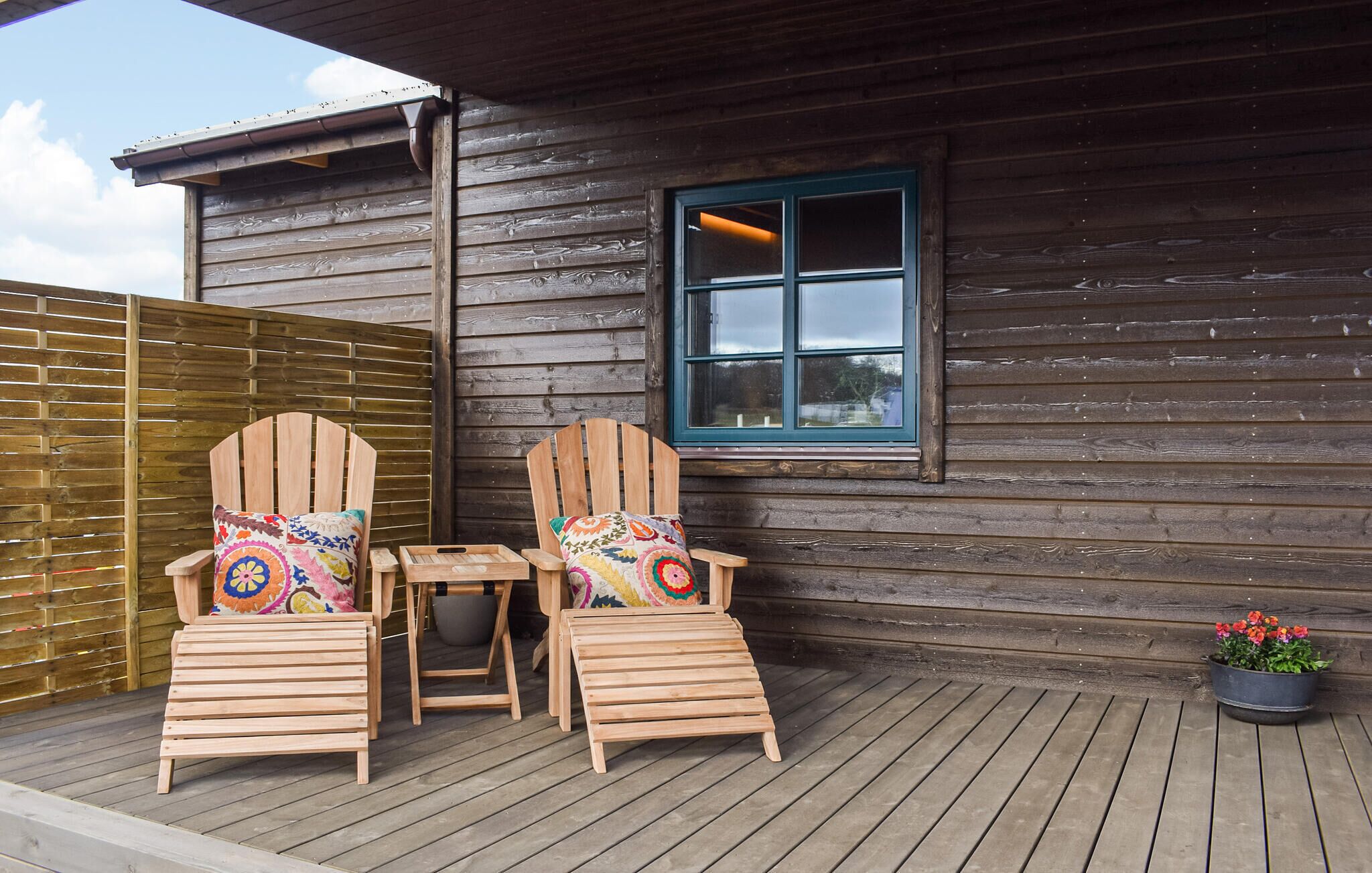 A wooden deck with two wooden chairs and a small table, a window with a blue frame, and a potted plant with flowers.