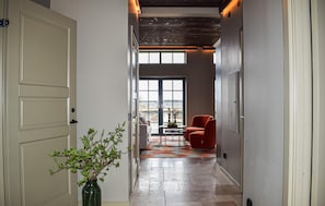 Un pasillo con una puerta, un jarrón con follaje verde, una silla roja y una mesa con un cuenco.