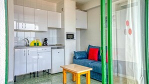 Cocina compacta con muebles blancos, microondas y un refrigerador pequeño. Hay una mesa de madera y un sofá azul con cojines rojos.