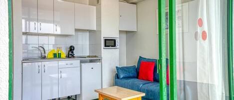 A compact kitchen with white cabinets, a microwave, and a small refrigerator. There's a wooden table and a blue sofa with red cushions.