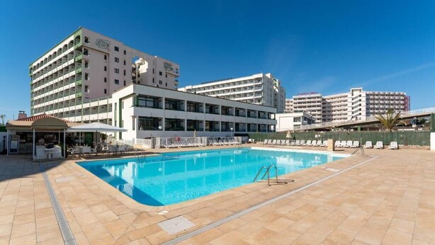 Un hotel con piscina, mobiliario exterior y varios edificios al fondo.