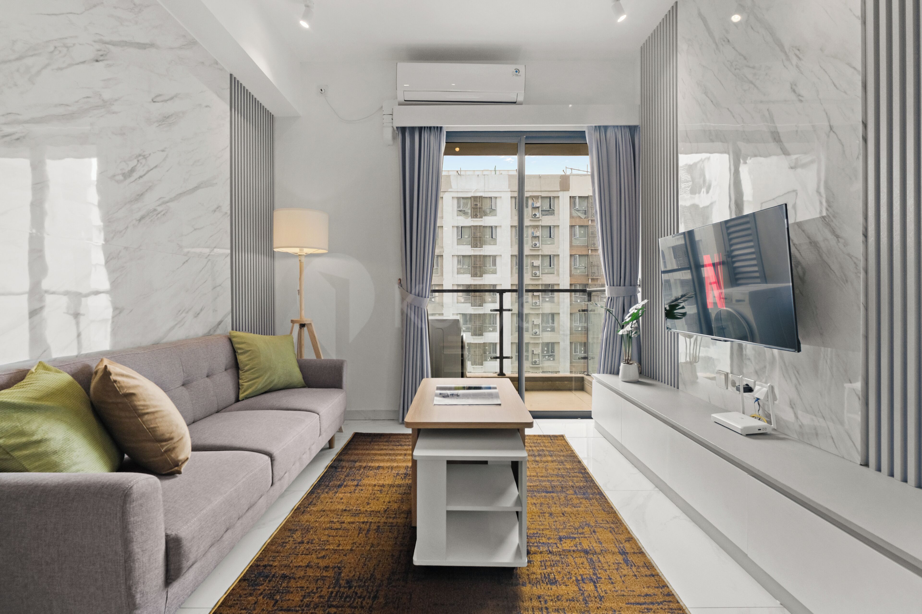 A modern hotel room with a grey sofa, a small table, a flat-screen TV, and a balcony with a view of the city.