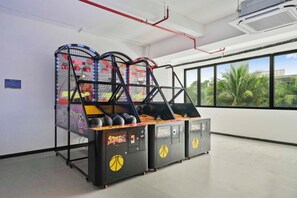 Eine Basketball-Spielhalle mit drei Automaten, einer digitalen Anzeigetafel und Blick auf Palmen durch große Fenster.
