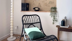 Ein Zimmer mit strukturierter Wand, ein schwarzer Rattan-Sessel mit grüner Kissenlehne, eine weiße Stehlampe, ein Holzregal mit Büchern und einem Wählscheibentelefon sowie eine hängende Pflanze.