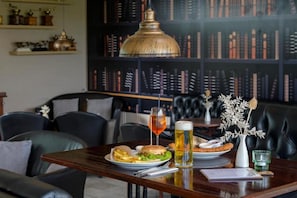 A dining table with a burger, fries, and a beer, set in a room with leather chairs and a bookshelf in the background.