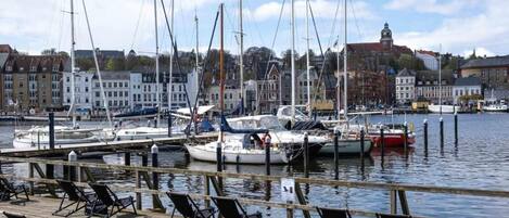 A marina with boats docked, a wooden pier with chairs, and buildings in the background.