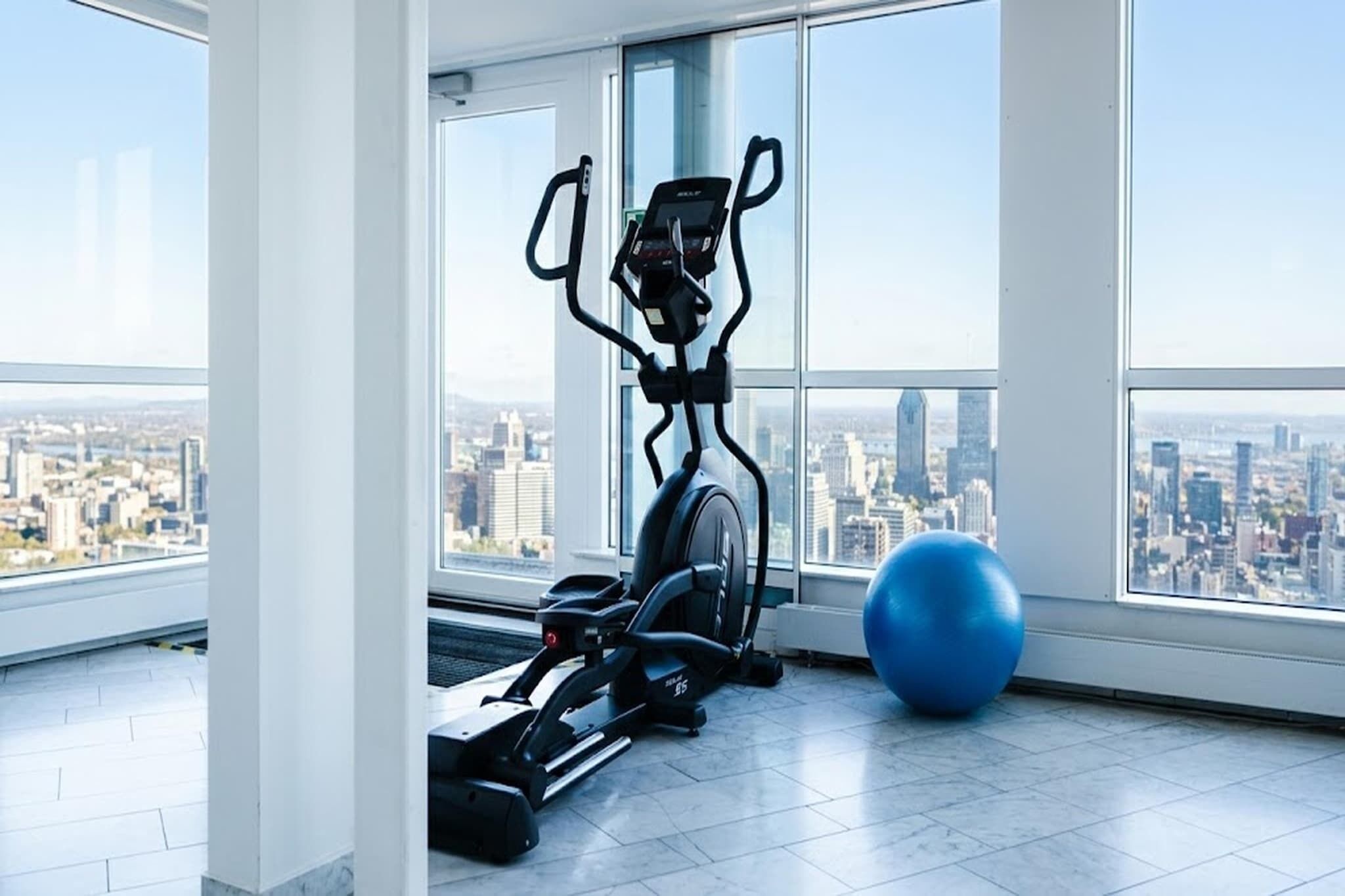 A high-rise room with a city view, an elliptical trainer, and a blue exercise ball.