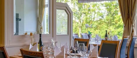 A dining area with tables set for a meal, chairs, and a view of greenery outside.