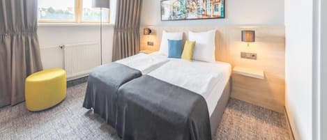 Habitación de hotel con dos camas, un reposapiés amarillo, una ventana con cortinas y un cuadro en la pared.
