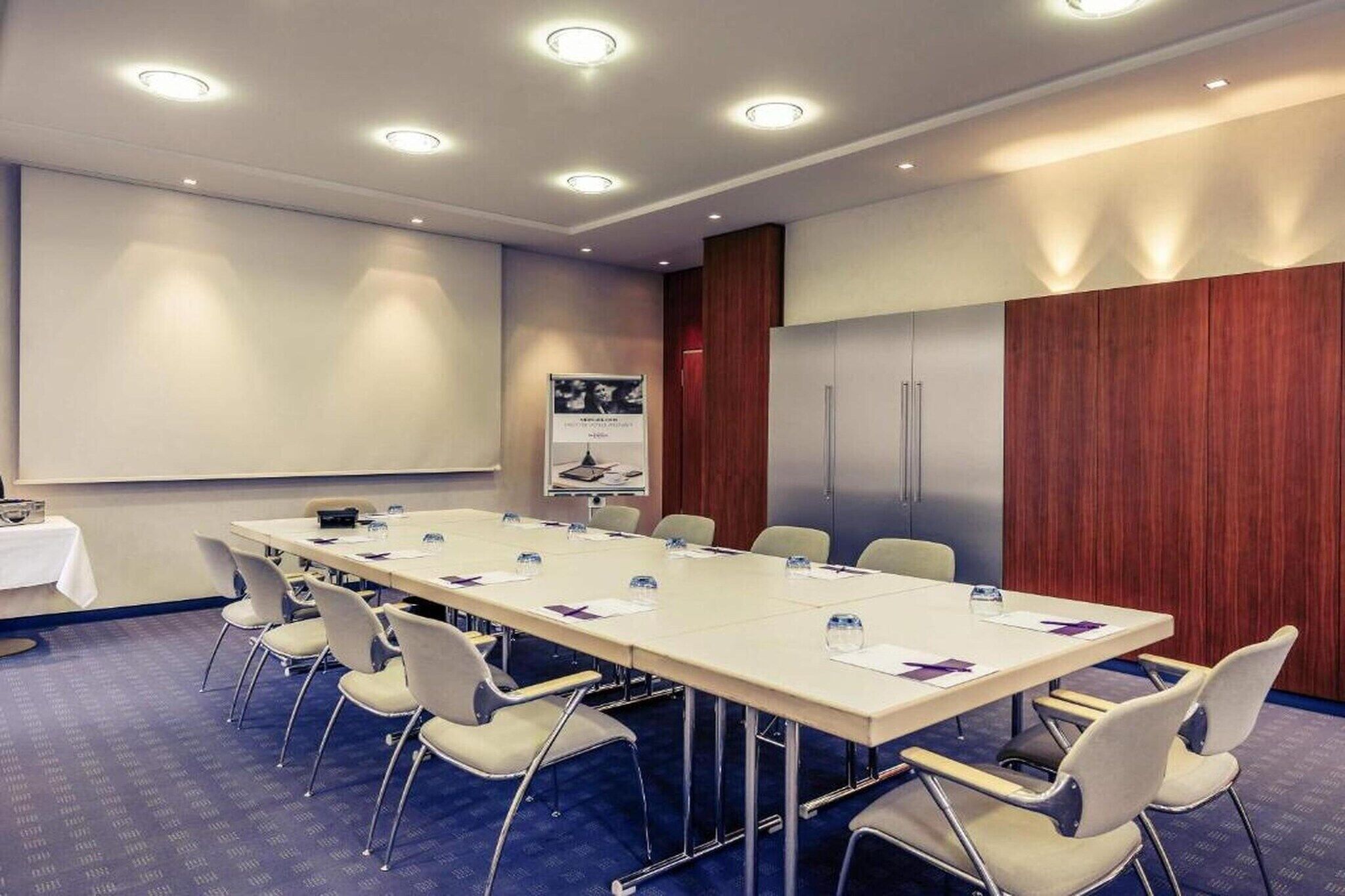 Una sala de conferencias con una mesa larga, sillas, una pantalla de proyector y un armario de madera.