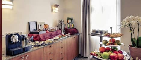 A hotel room with a kitchenette, a coffee machine, a fruit platter, and a tiered dessert stand.