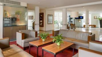 Eine moderne Hotelhalle mit Ledersofas, Holztischen und einer Glastrennwand, die den Sitzbereich von der Küche abteilt.