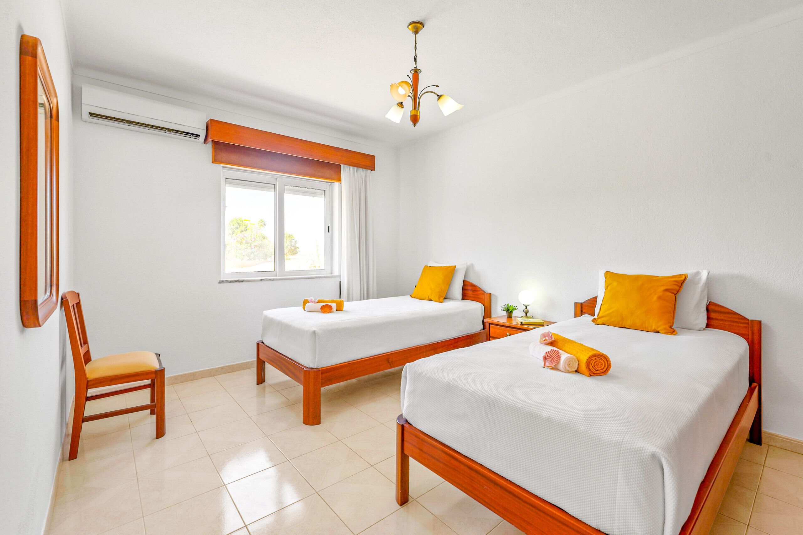 A hotel room with two single beds, a wooden chair, a window with curtains, a ceiling light, and a wall-mounted air conditioner.