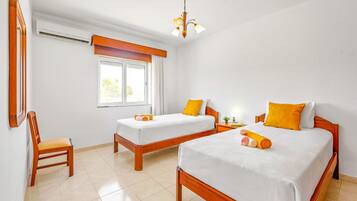 A hotel room with two single beds, a wooden chair, a window with curtains, a ceiling light, and a wall-mounted air conditioner.