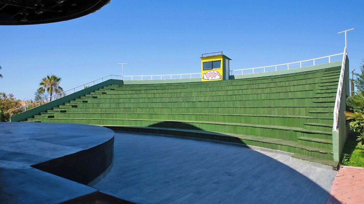 Een stadion met tribuneaanstellingen, een centrale gele constructie en een helderblauwe lucht.