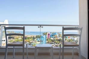Un balcón con dos sillas blancas y una mesa, con vista a una playa y un parque acuático.
