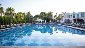 Une piscine à motifs à carreaux, entourée de chaises longues et de parasols, avec des palmiers et un bâtiment en arrière-plan.