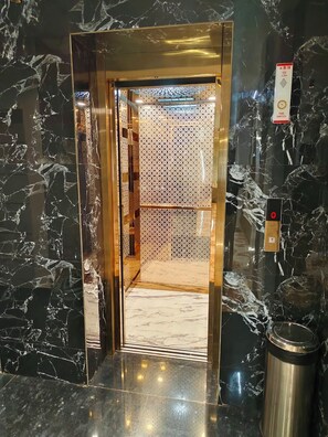 An elevator with a reflective metallic interior, a fire alarm switch, and a trash bin.