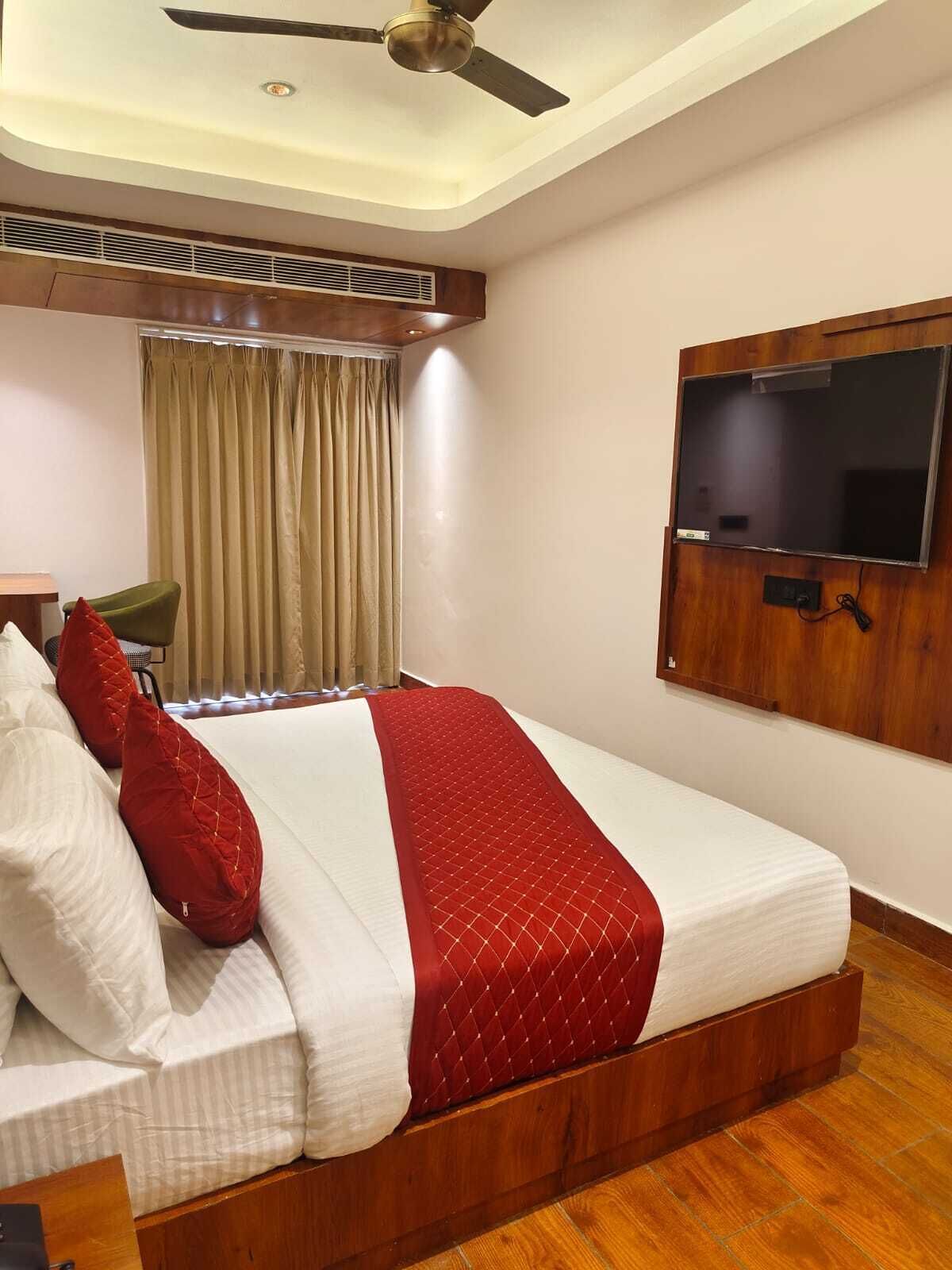 A hotel room with a bed, a television mounted on the wall, a ceiling fan, and a window with curtains.