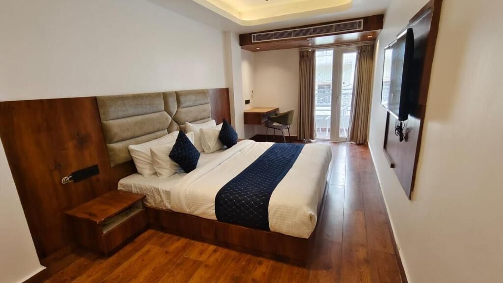 Ein Hotelzimmer mit einem großen Bett, einem Schreibtisch, einem Stuhl, einem Fernseher und einem Balkon mit Vorhängen.