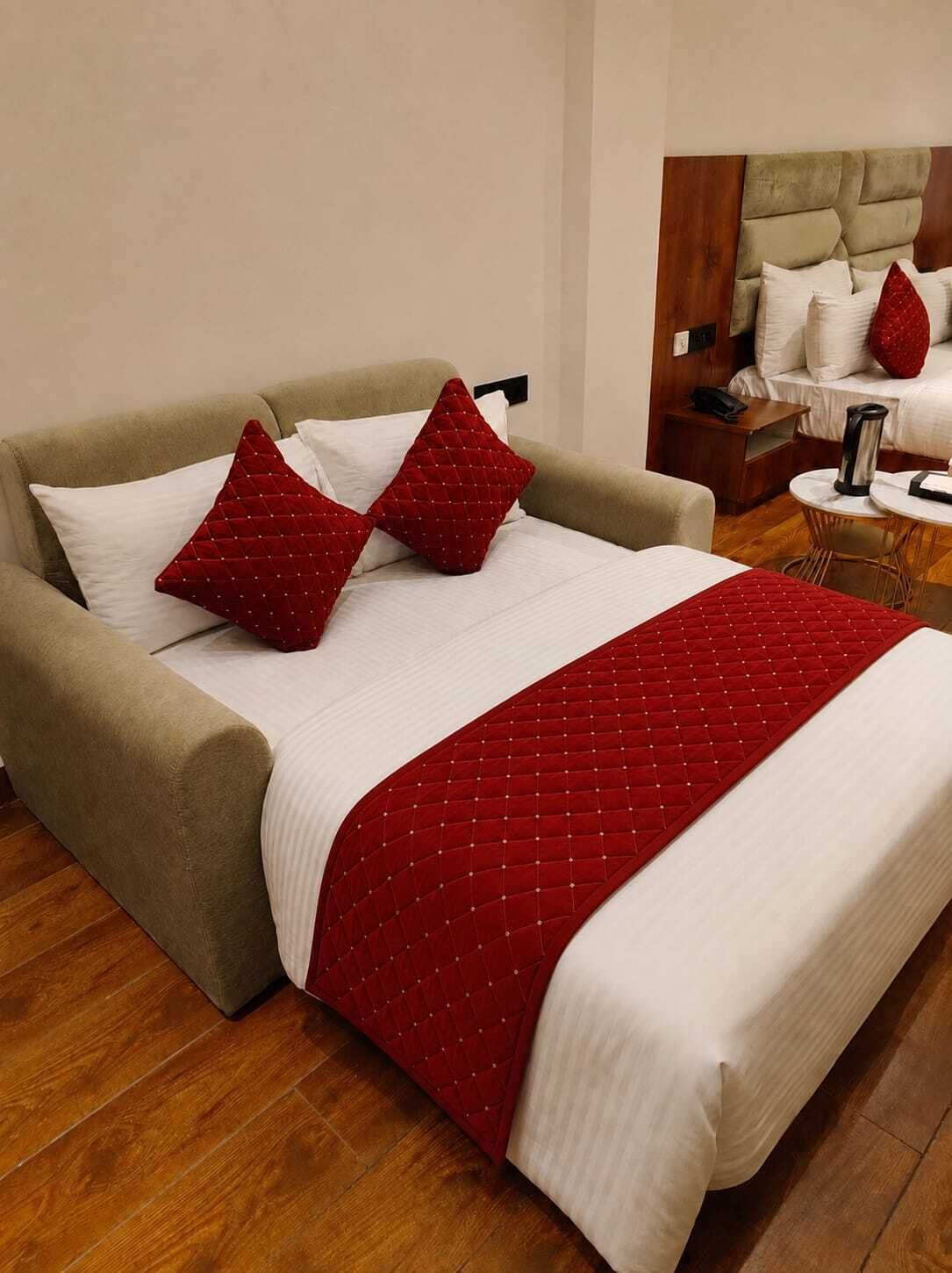 A bed with a quilted red blanket and two pillows, a wooden floor, and a nightstand with a lamp.