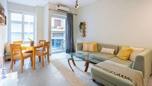 Ein modernes Wohnzimmer mit einem Glastisch, einer Couch mit Kissen, einem Essbereich mit Stühlen und einem sichtbaren Balkon durch das Fenster.
