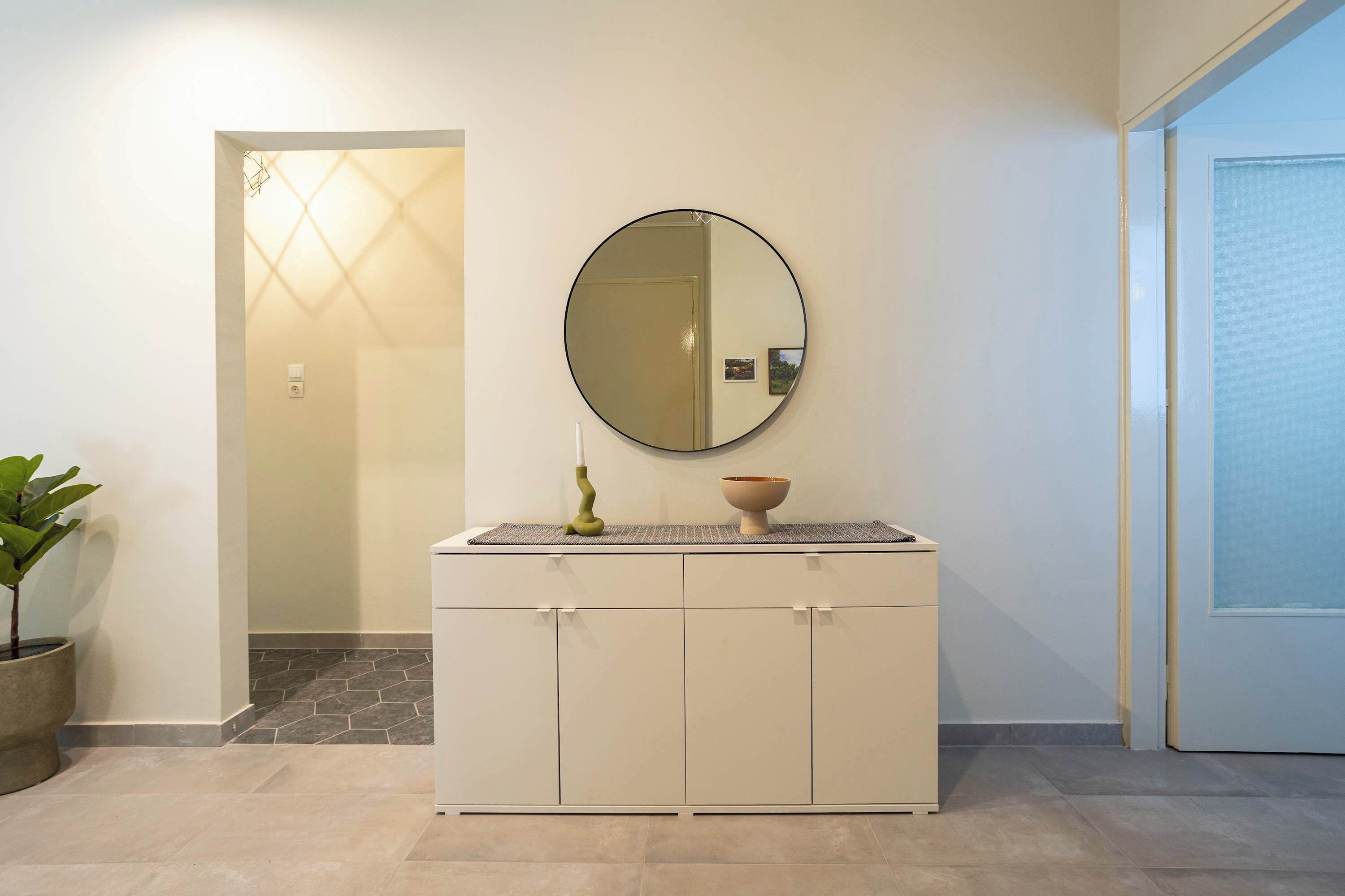 A hallway with a white cabinet, a round mirror, a potted plant, and a door with a frosted glass panel.