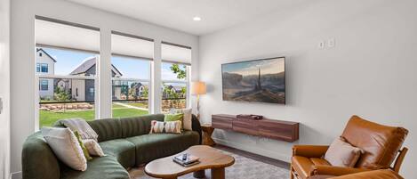 A spacious living room with a sectional sofa, a leather armchair, a coffee table, and a flat-screen TV mounted on the wall.