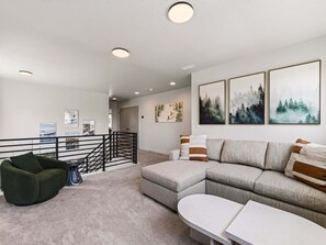 A modern living room with a grey sofa, a green armchair, and a white coffee table. There are three framed artworks on the wall, a staircase with a black railing, and a door leading to another room. - New! Deer Valley Close, 4BR 3.5BA, Sleeps 11, BBQ (Heber City)