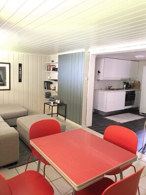 Un espacio habitable compacto con kitchenette, un comedor con sillas rojas, un sofá y una estantería.