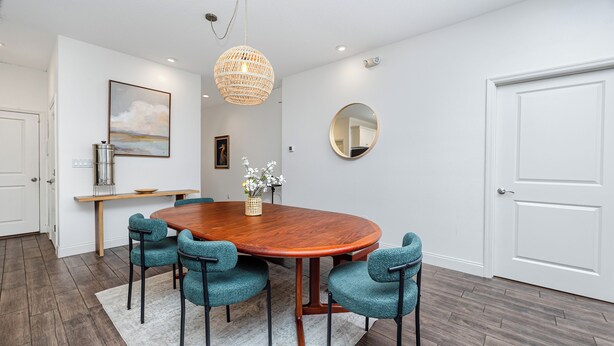 Un coin repas comprenant une table en bois, quatre chaises de couleur turquoise, un luminaire suspendu en fibres tressées, un miroir rond et une porte blanche.