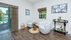 Un salon moderne avec une porte vitrée donnant sur une piscine, deux fauteuils gris, une petite table en bois, une table d’appoint en métal noir agrémentée d’objets décoratifs et un tableau abstrait encadré.