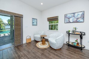 Ein modernes Wohnzimmer mit Glastür zum Pool, zwei graue Sessel, ein kleiner Holztisch, ein schwarzer Metall-Beistelltisch mit Dekoration und ein gerahmtes abstraktes Gemälde.