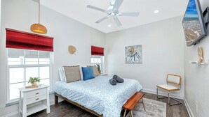 Une chambre à coucher avec un lit, une chaise, un bureau, une fenêtre avec des stores rouges, un ventilateur de plafond et un tableau accroché au mur.