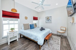 Ein Schlafzimmer mit einem Bett, einem Sessel, einem Schreibtisch, einem Fenster mit roten Jalousien, einem Deckenventilator und einem Bild an der Wand.