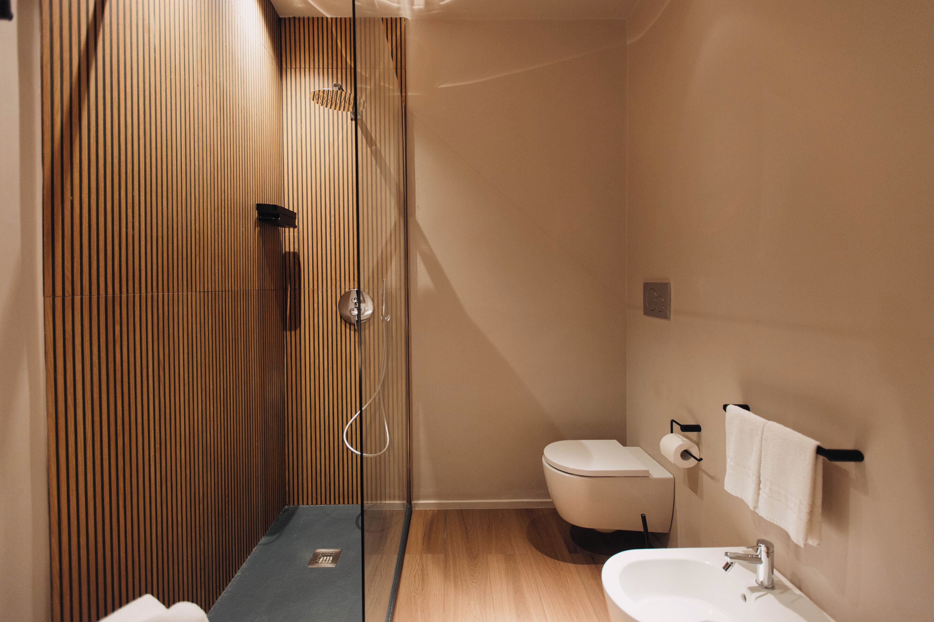 A modern bathroom with a wooden panel wall, a glass shower enclosure, a white toilet, and a sink with a towel hanging on the wall.
