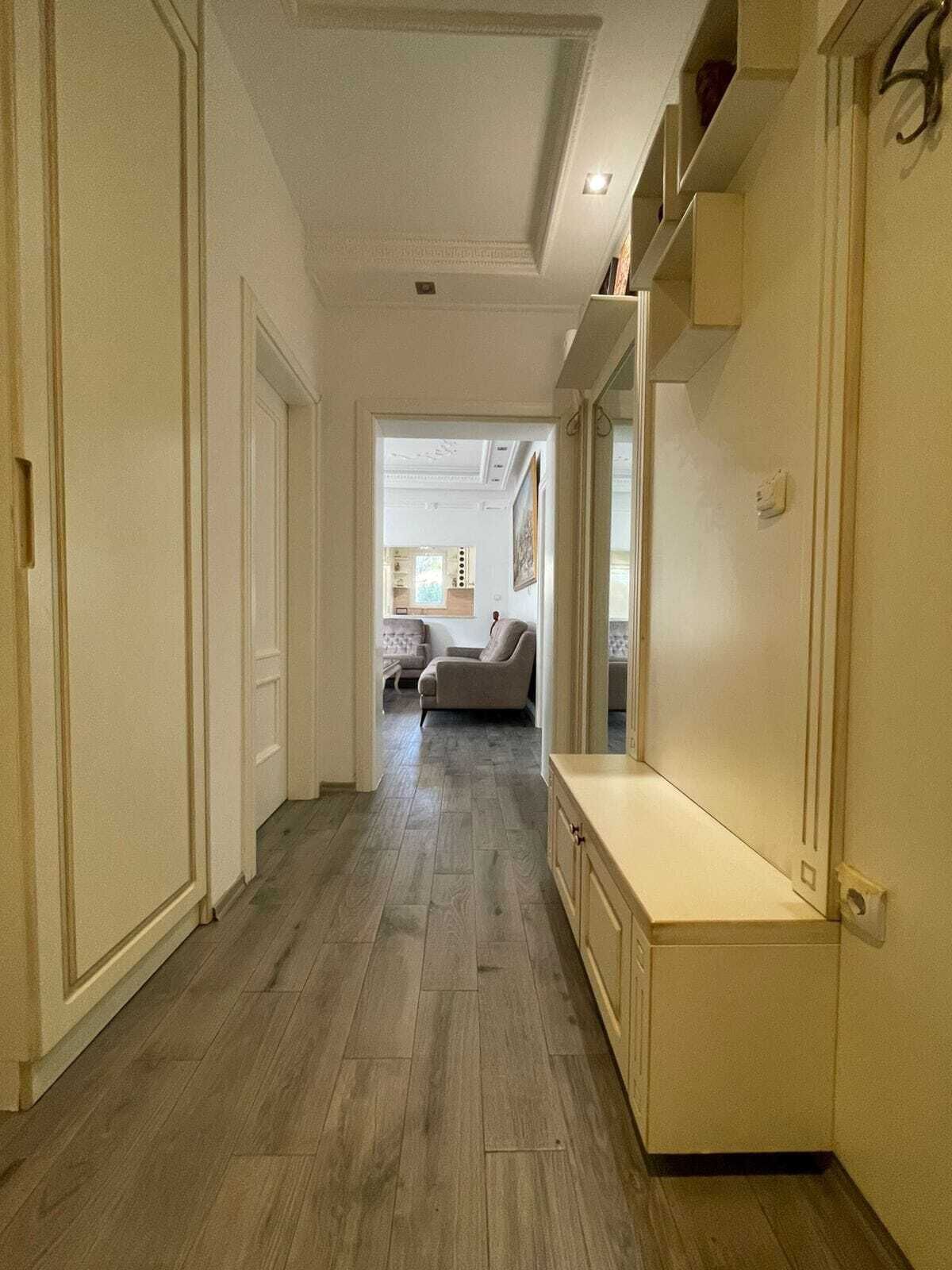 A hallway with a wooden floor, white walls, and a door leading to a room with a bed and a window.