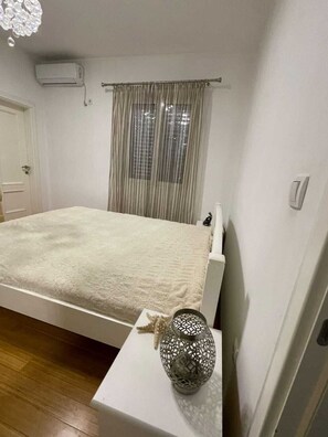 A bedroom with a bed, a window with curtains, an air conditioning unit, and a decorative vase on a nightstand. - Modern Classic 2-Bedroom Apartment in Donja Lastva (Tivat)