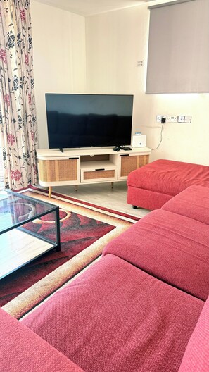 Ein Wohnzimmer mit Fernseher, einem roten Sofa, einem Glastisch und einem gemusterten Teppich.