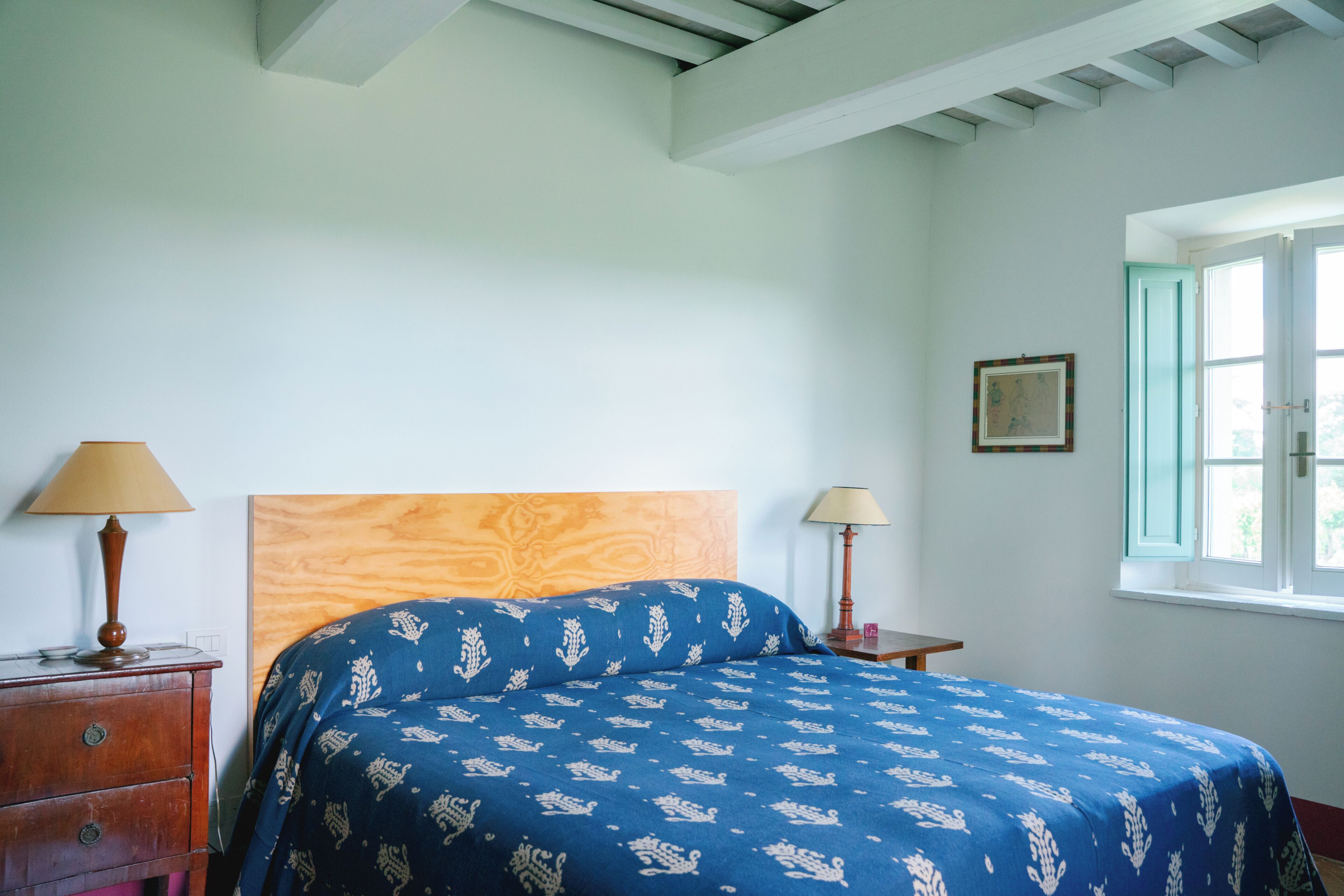 Une chambre à coucher avec une tête de lit en bois, un lit recouvert d’une couvre-lit à motifs bleus, deux tables de chevet avec des lampes et une fenêtre donnant sur un paysage agréable.